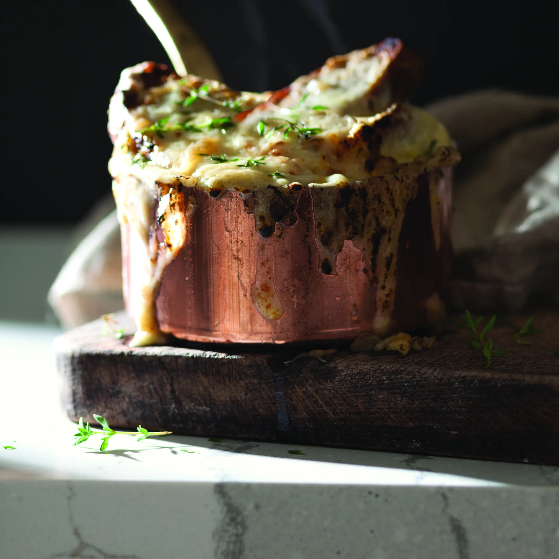 A steaming copper pot of baked onion soup resting on a thick wooden board on a white veined quartz countertop