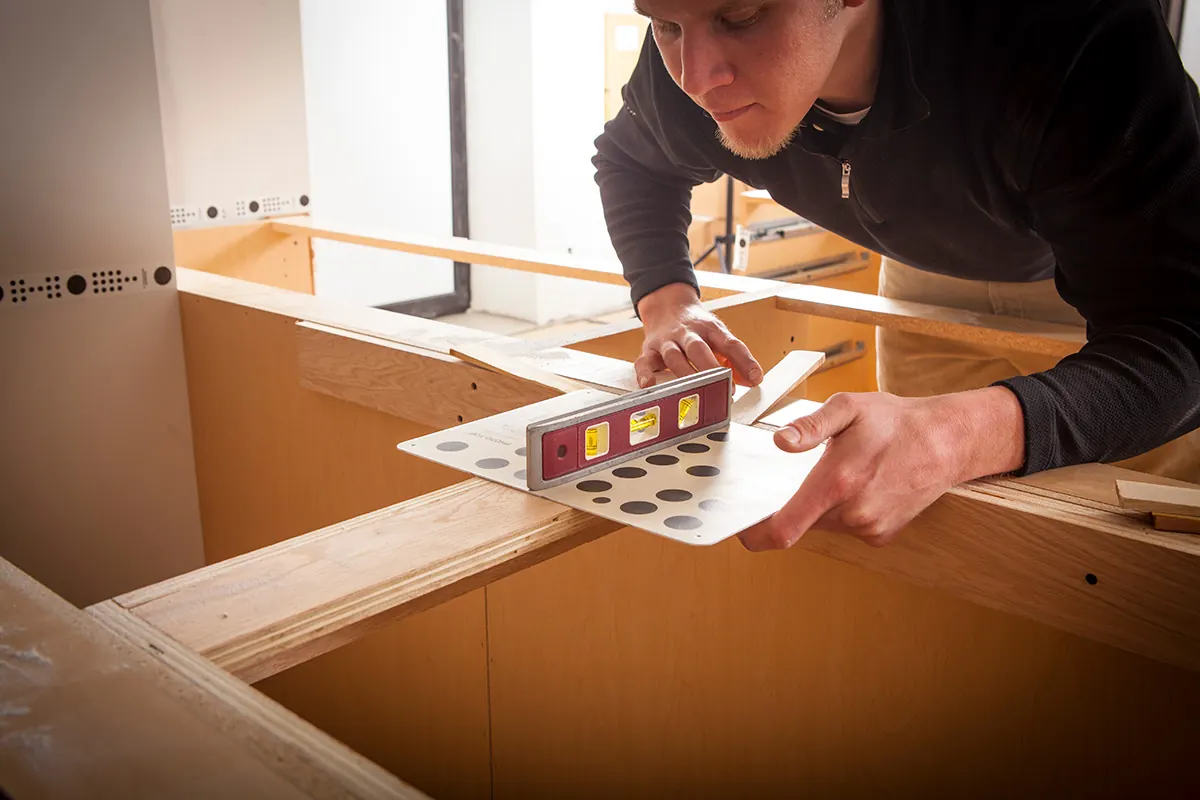 A man holding a small red level on a dotted measuring target across the top rails of bare base cabinets
