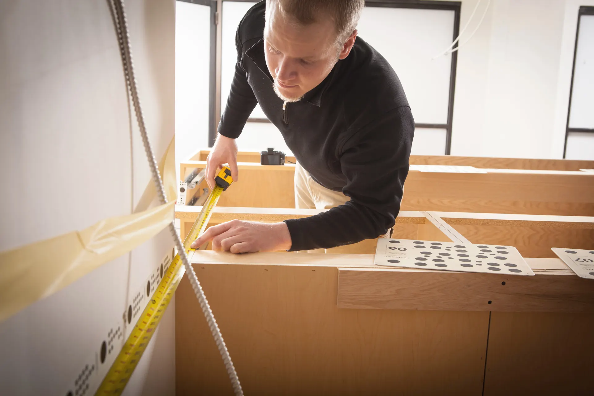 A man stretching a tape measure across bare wood base cabinets that have no countertop on them yet