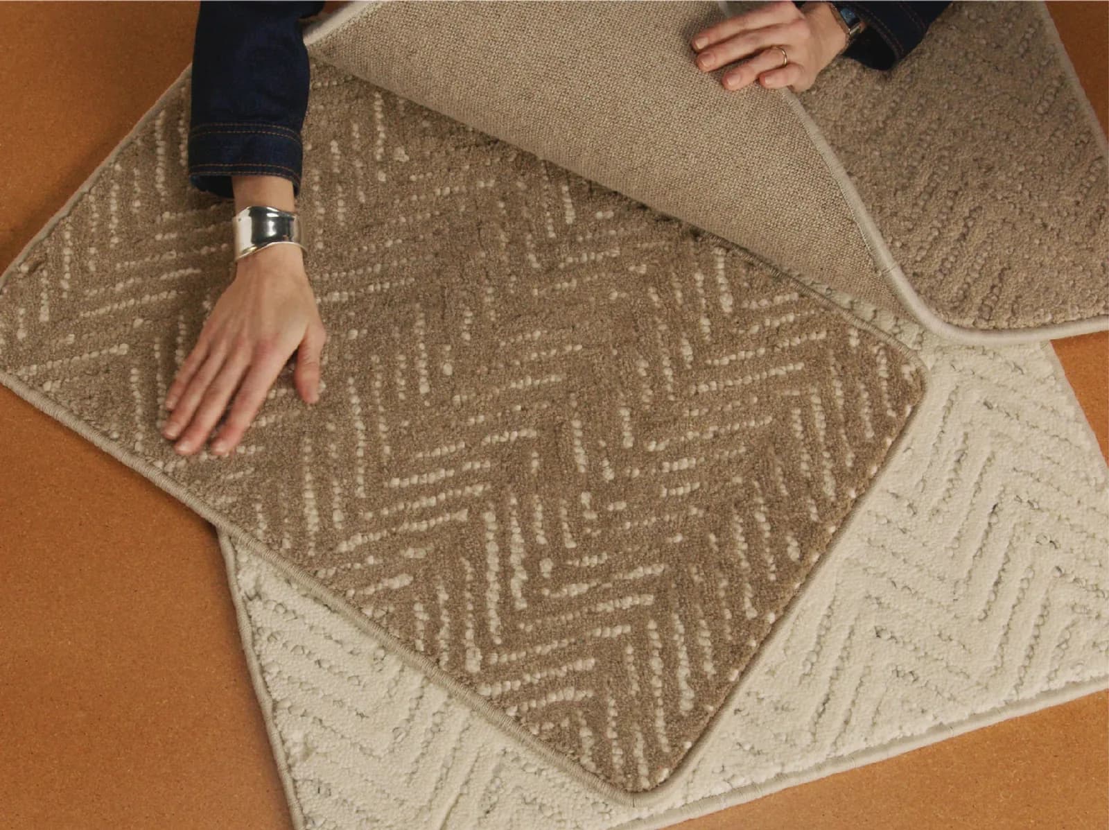 Two carpet swatches laid on a rust-orange surface, one folded back over the other so the pile face, the fold and the cut edge are all visible at once, with a person's hands holding them flat