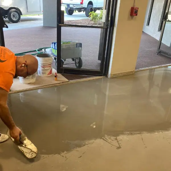 An installer spreads self-leveling underlayment across a bare slab, with a bucket of First Coat primer beside him.