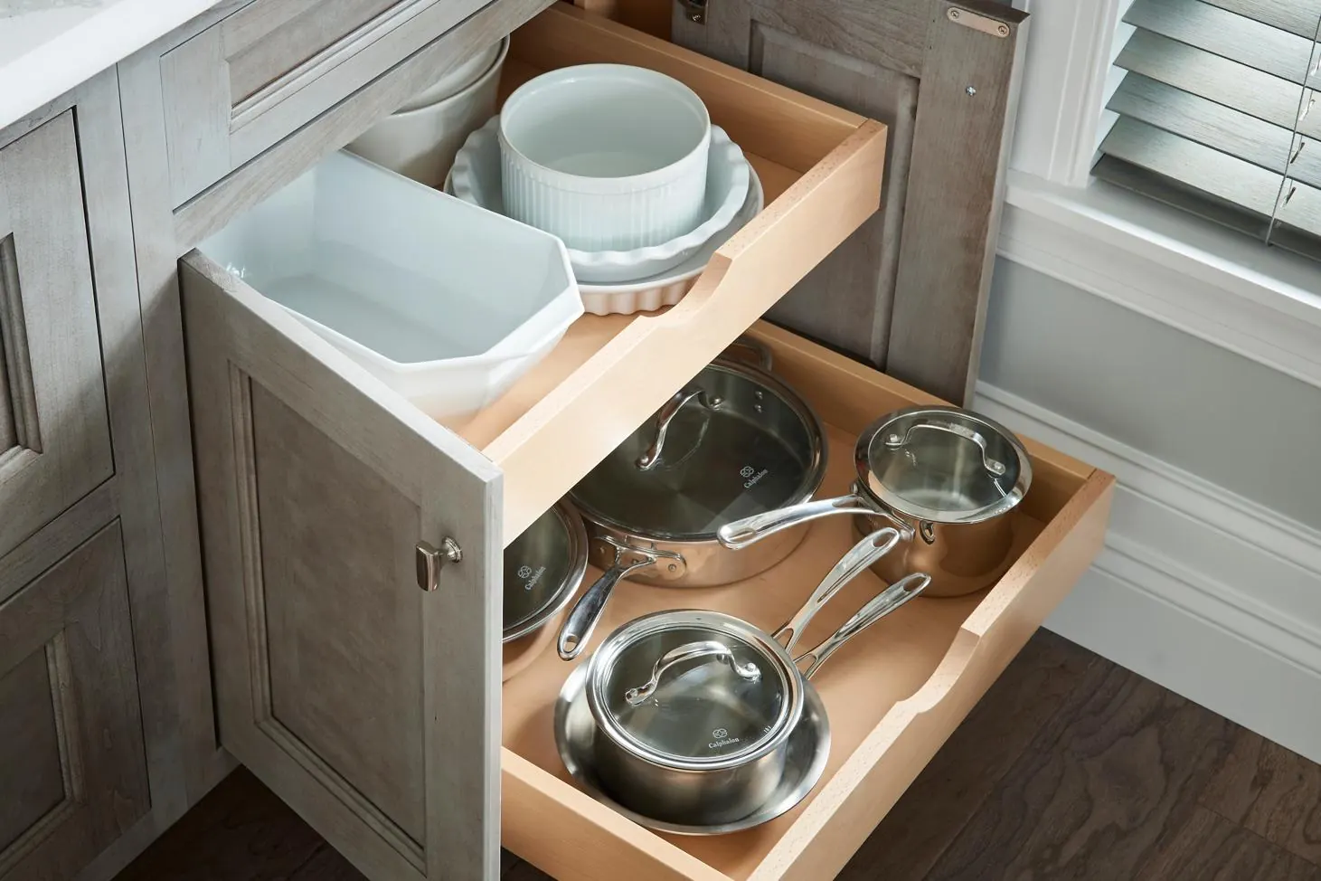 Open base cabinet door with two wood roll-out trays holding pots, pans and white baking dishes