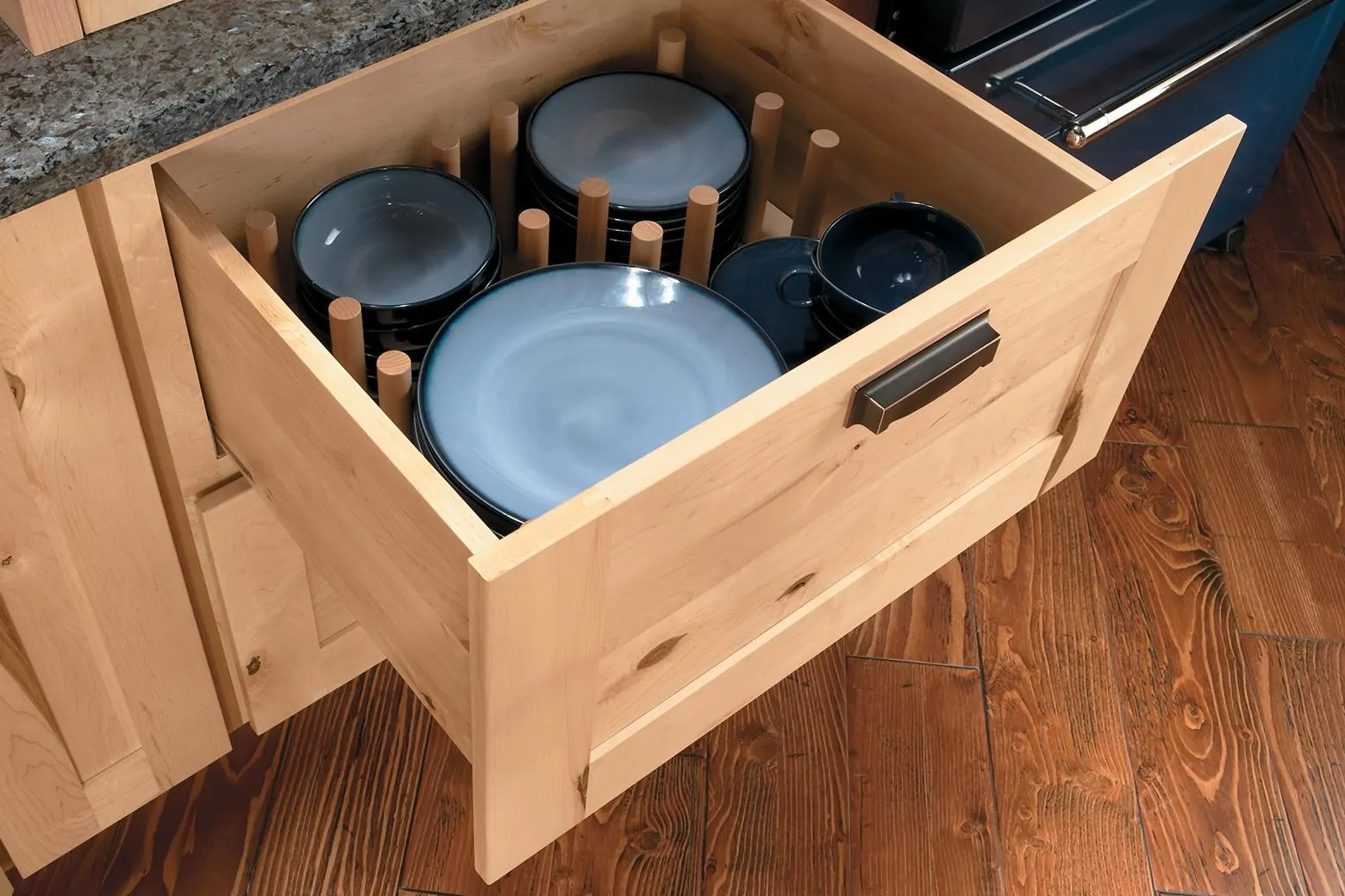 Deep wood base cabinet drawer pulled open with stacked blue plates and bowls held by wooden pegs