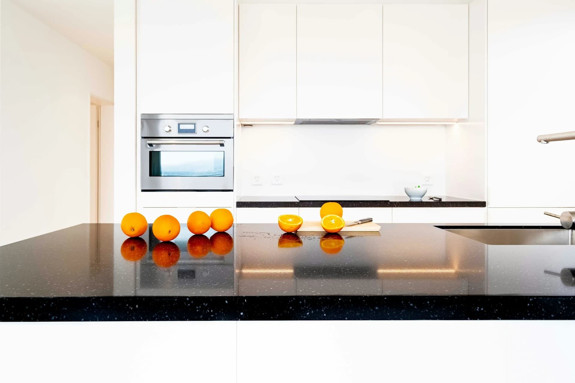 Black speckled quartz countertop on a kitchen island. White cabinets and a wall oven sit behind. Oranges rest on a cutting board.