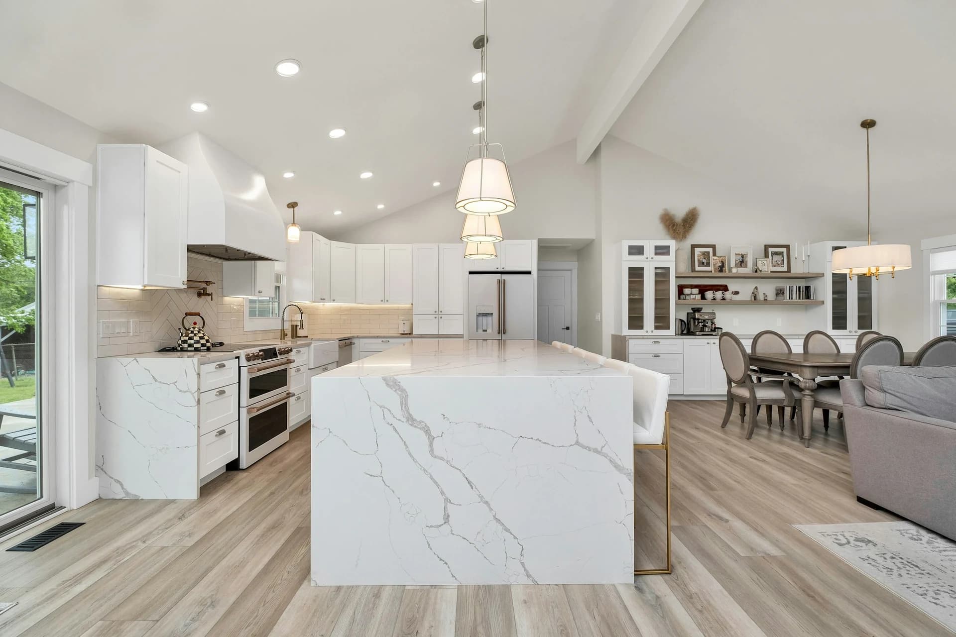 A white waterfall island with bold gray veining anchors a bright open kitchen with white cabinets and light plank floors.