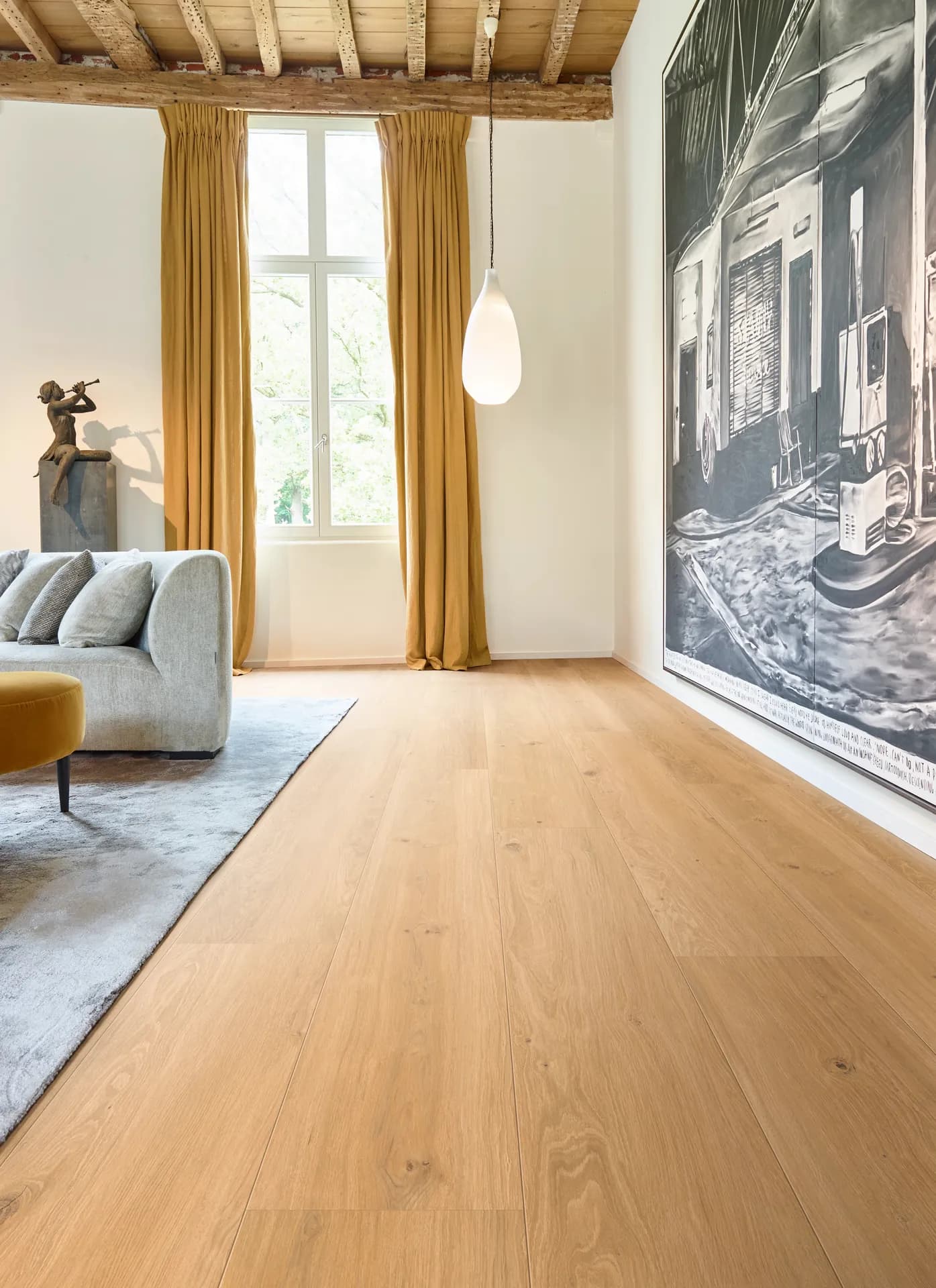 Light oak wide plank hardwood in a bright living room with a gray sofa and gold curtains. Hardwood flooring for Bartow, FL homes