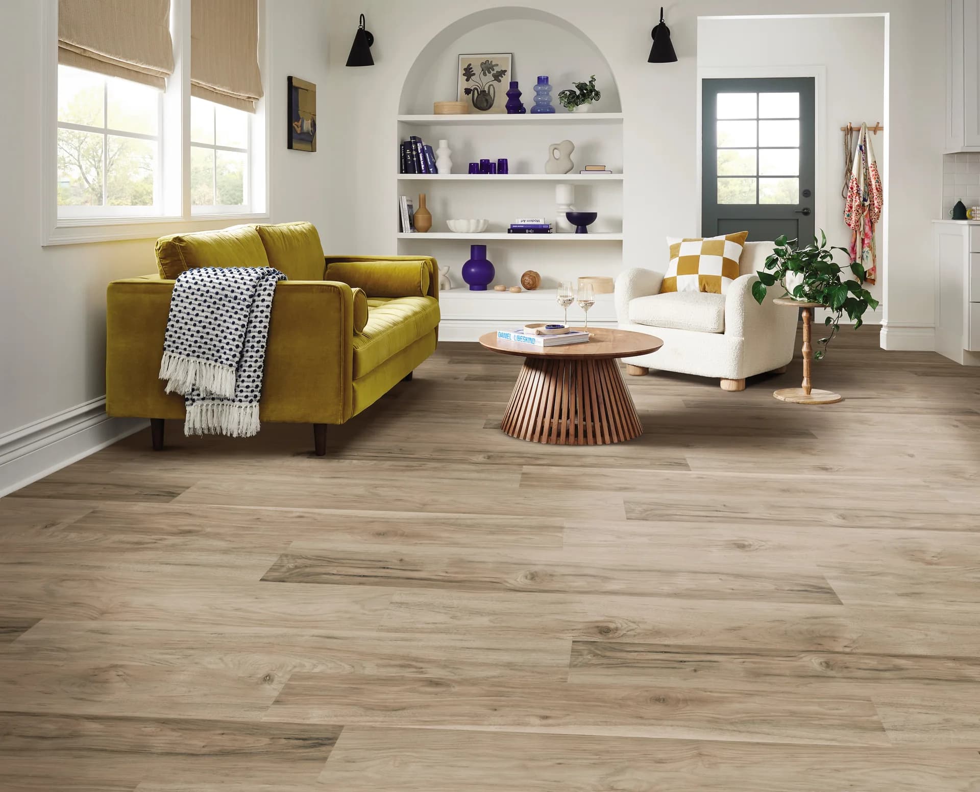 Shaw Floors Ignite luxury vinyl plank in Iron Fog, a warm gray-brown wood look, in a white living room with a mustard velvet sofa.