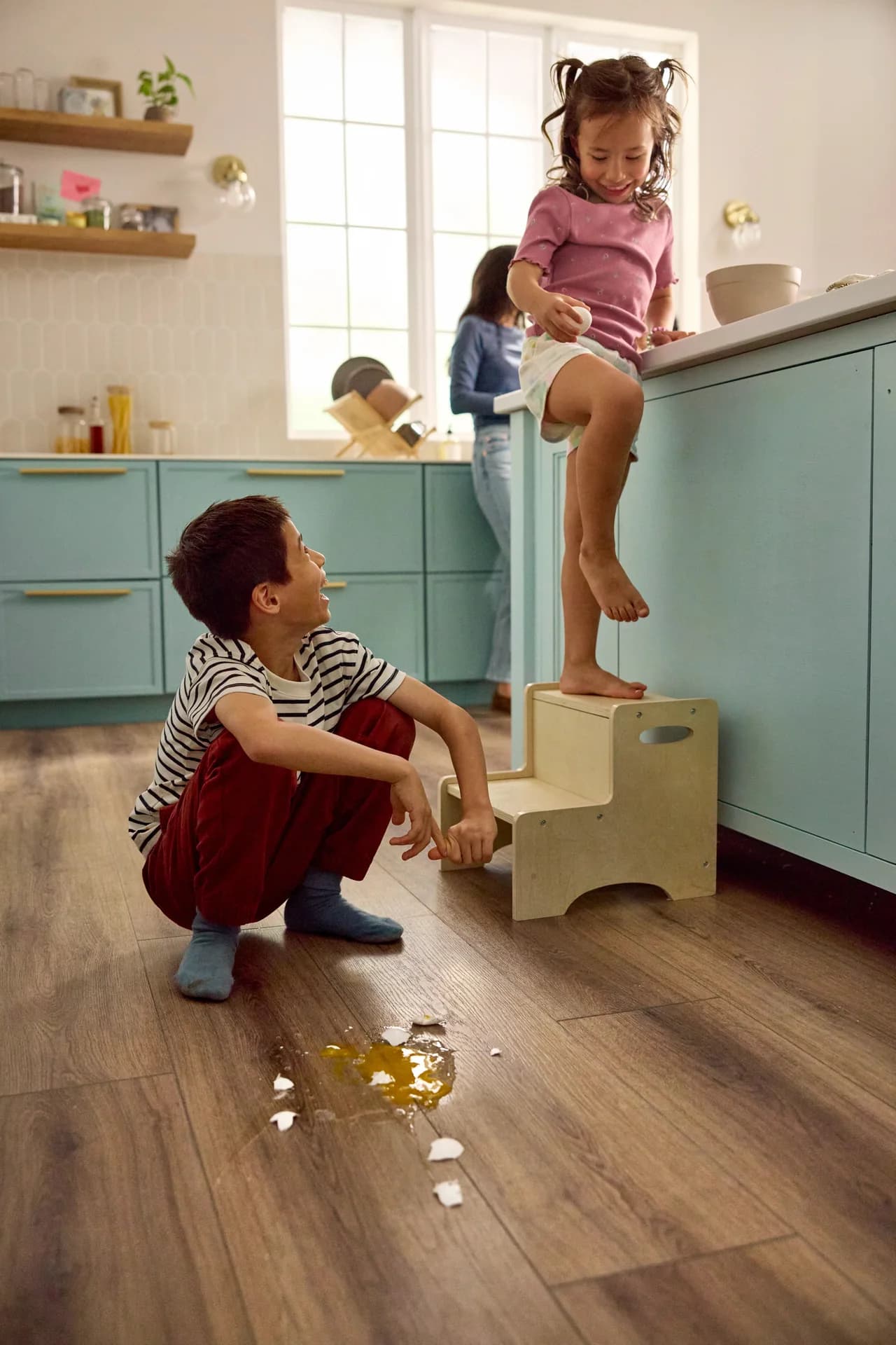 Shaw Floors Pet Perfect Briard vinyl plank in Coffee Oak runs across a kitchen floor with teal cabinets. Two kids sit and stand near a spilled egg.
