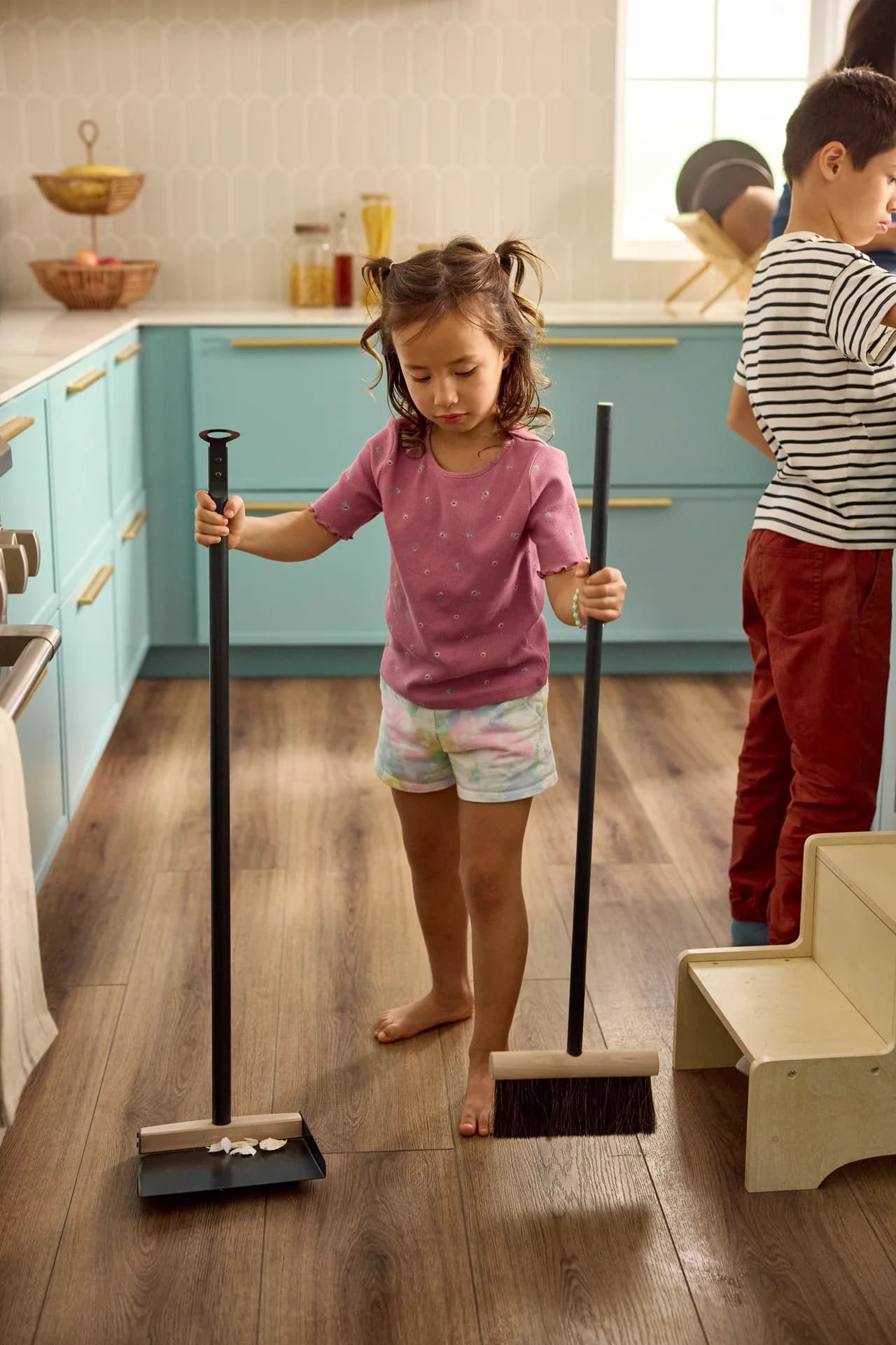 Shaw Floors Pet Perfect Briard luxury vinyl plank in Coffee Oak runs through a kitchen with teal cabinets. A barefoot girl sweeps bits into a dustpan.