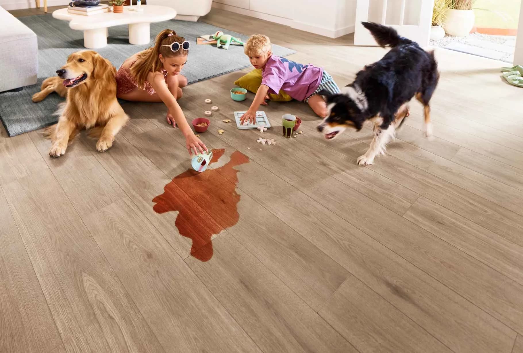 Two dogs and two kids on Shaw Cattitude vinyl plank in Fika, a light greige wood look, with a dark drink spilled across the floor.