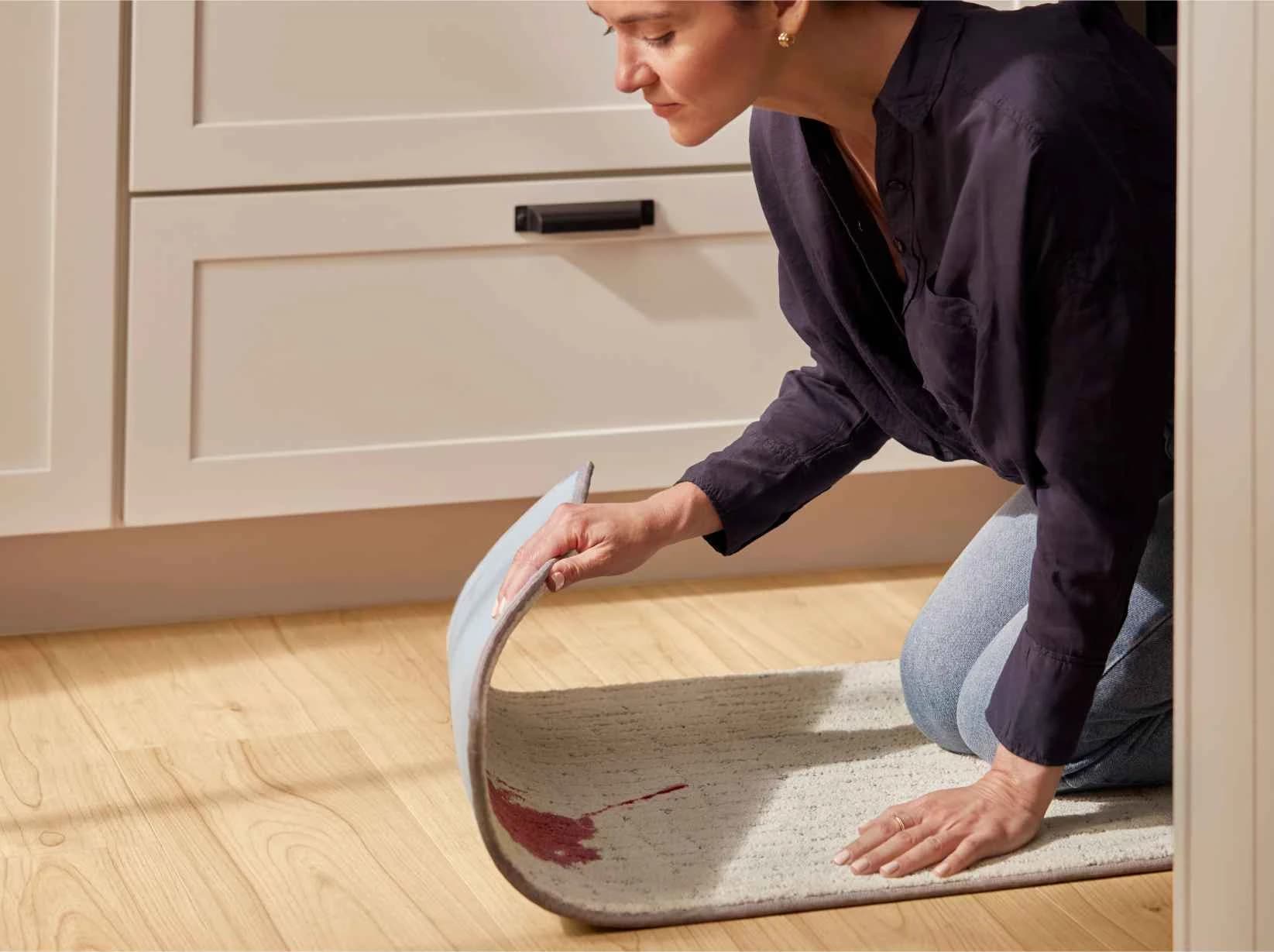 Woman lifts the edge of a cream Shaw Floors carpet area rug on a light wood-look plank floor, a red spill on the rug and the wood floor beneath it dry.