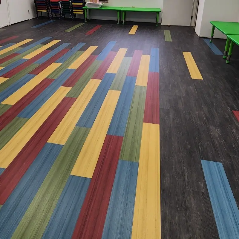 Edge of the color rectangle blending into the dark LVP main floor with stacked kid chairs and tables along the back wall