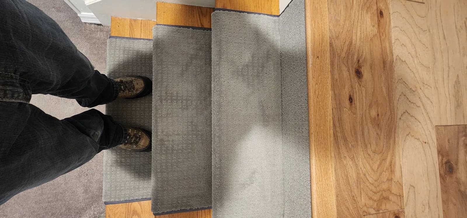 Bottom step where the bound nylon runner meets a wide-plank hardwood floor, an installer standing on the runner